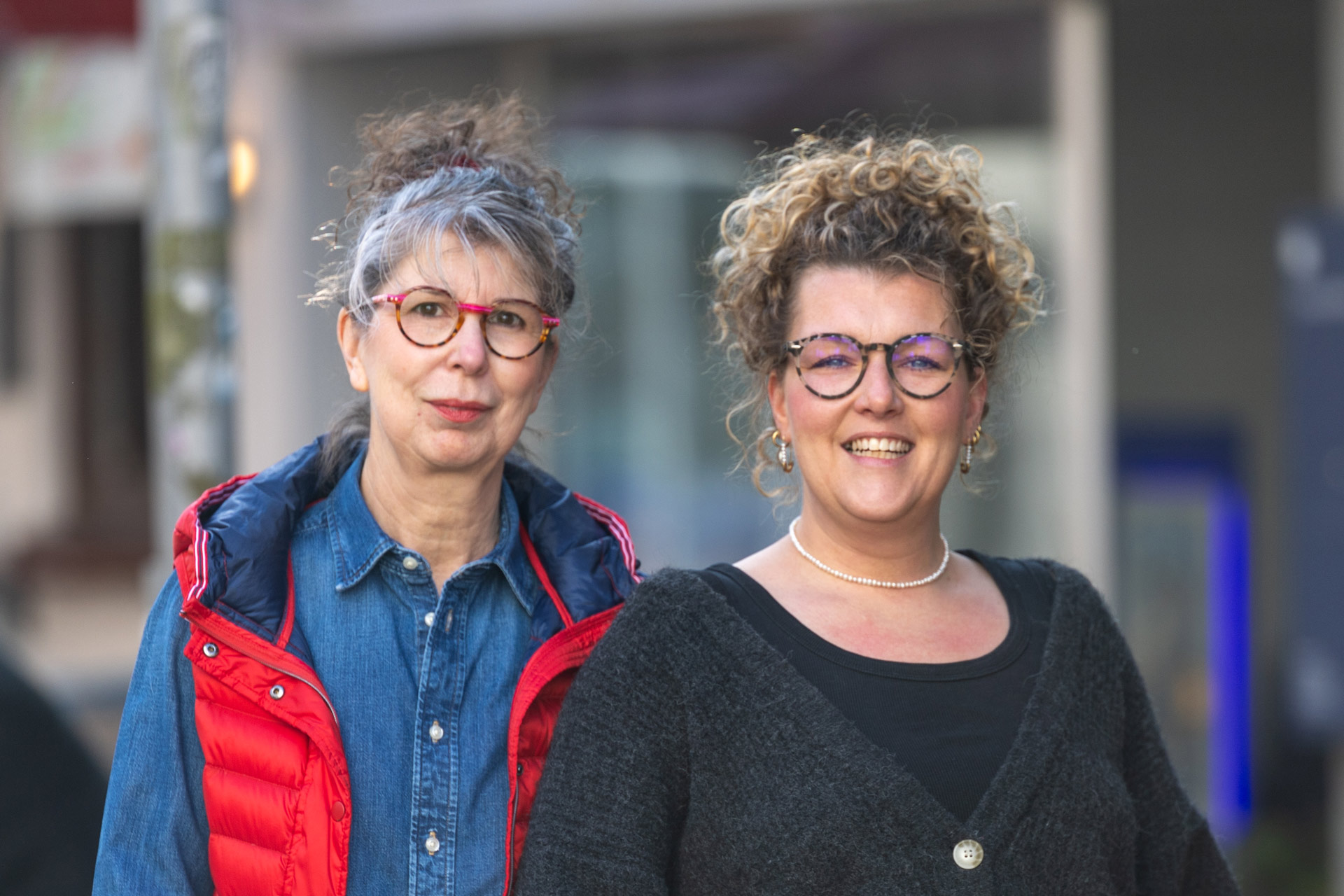 Zwei lächelnde Frauen mit Brille und lockigen Haaren stehen draußen nebeneinander